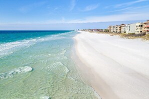 A beachfront with clear shallow waters, a sandy beach, and a row of multi-story buildings with balconies.