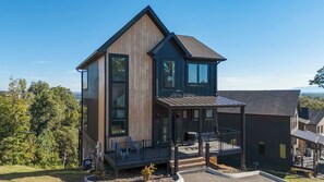 A modern two-story house with a large balcony, a covered outdoor seating area, and a view of the surrounding landscape. - Heated Indoor Pool, Luxury Cabin, Near Dollywood (Sevierville)