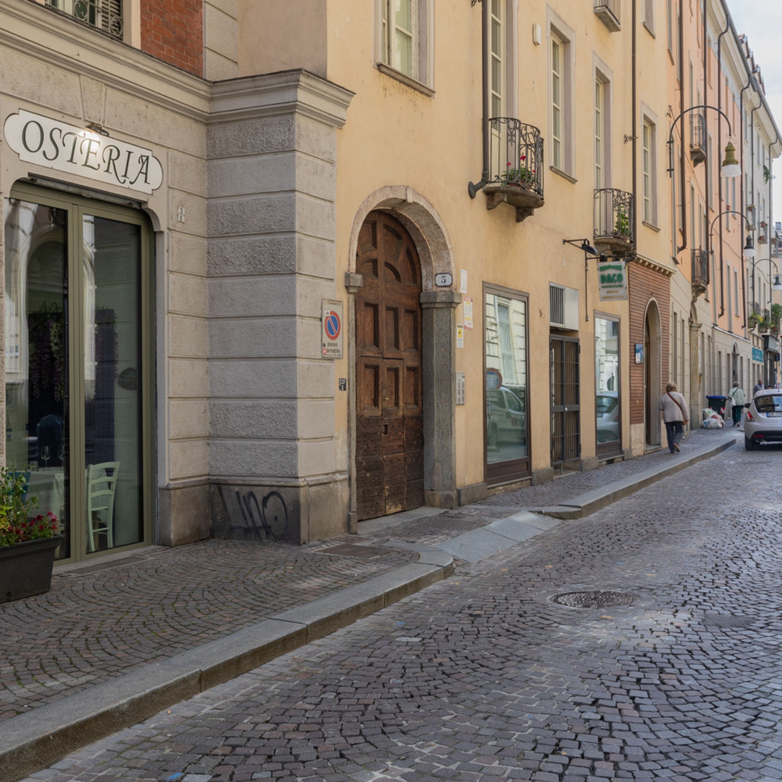 A street with cobblestone pavement, a building with a sign reading "OSTERIA", and a closed wooden door.