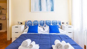 A hotel room with a bed, two bedside tables, a headboard with a blue and white pattern, a painting on the wall, and a window with curtains. - Casa Vacanze Nostos Alabardieri by Napoliapartments (Naples)