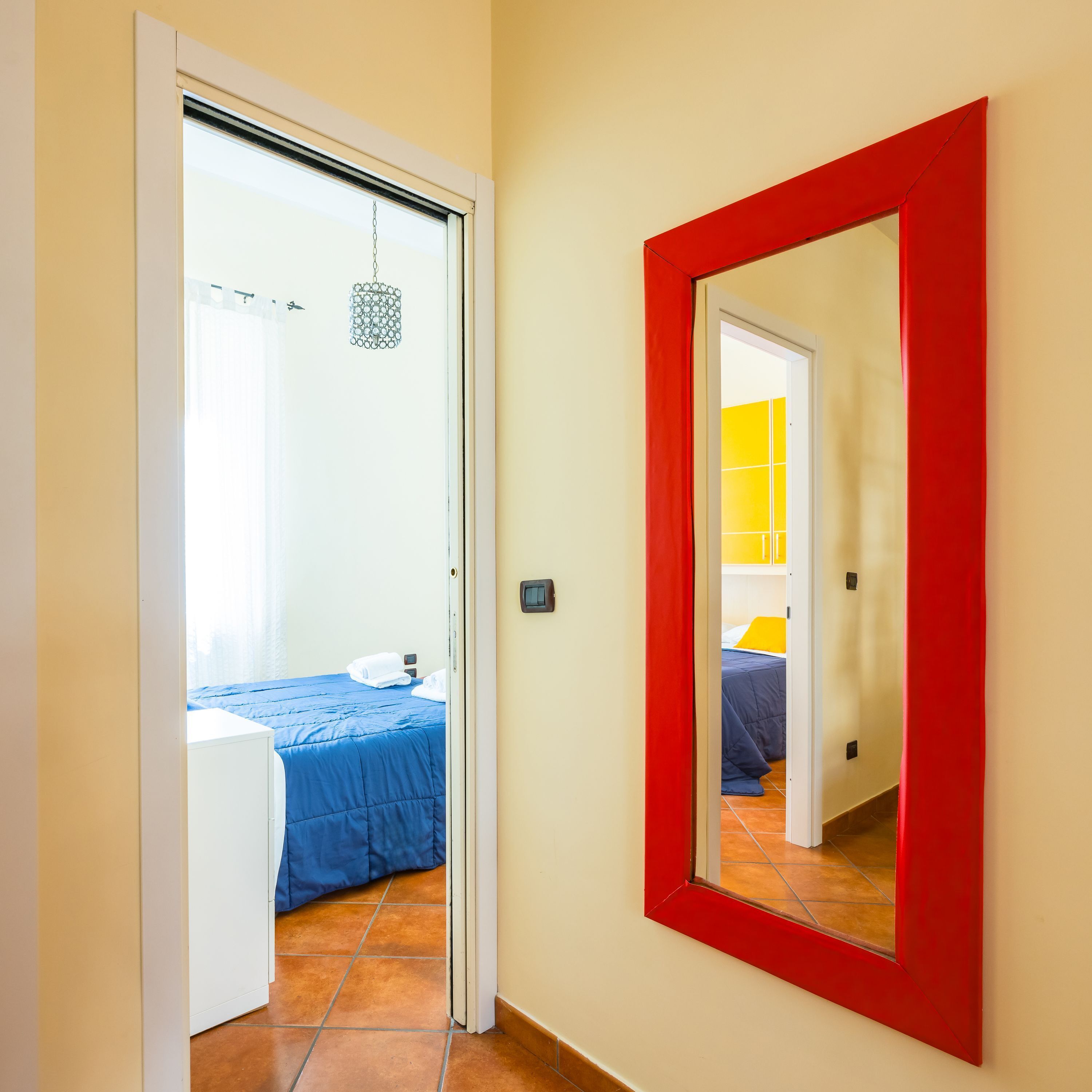 A hallway leading to a bedroom with a blue bedspread, a red-framed mirror, and a hanging light fixture.