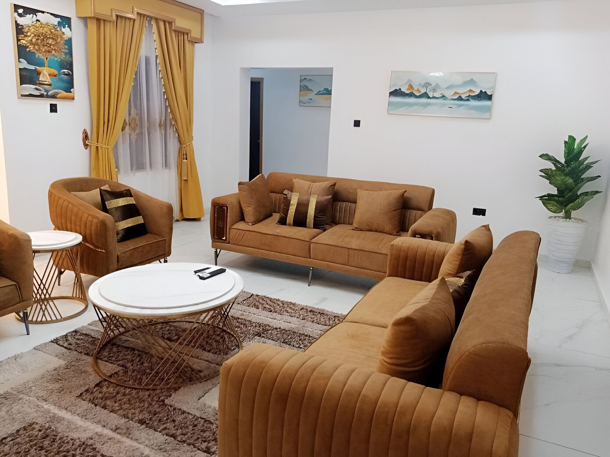 A living room with a brown sofa, a round coffee table, and a small side table. There are two paintings on the walls and a potted plant in the corner.