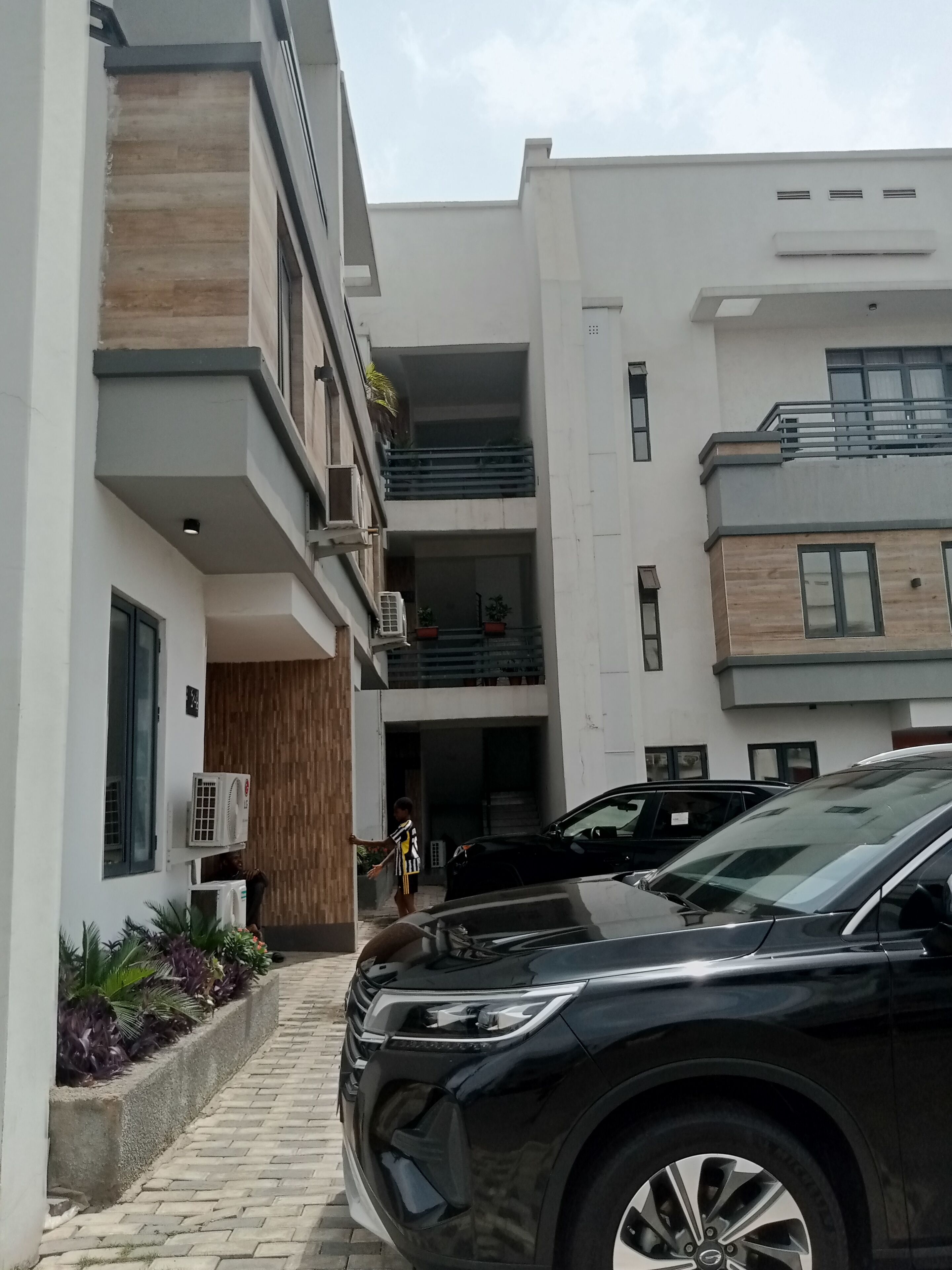 Um edifício moderno com entrada pavimentada, um SUV preto estacionado na frente e uma pessoa caminhando em direção à entrada.