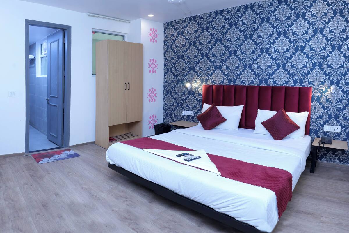 A hotel room with a bed, a desk with a chair, a wardrobe, and a bathroom visible through an open door.