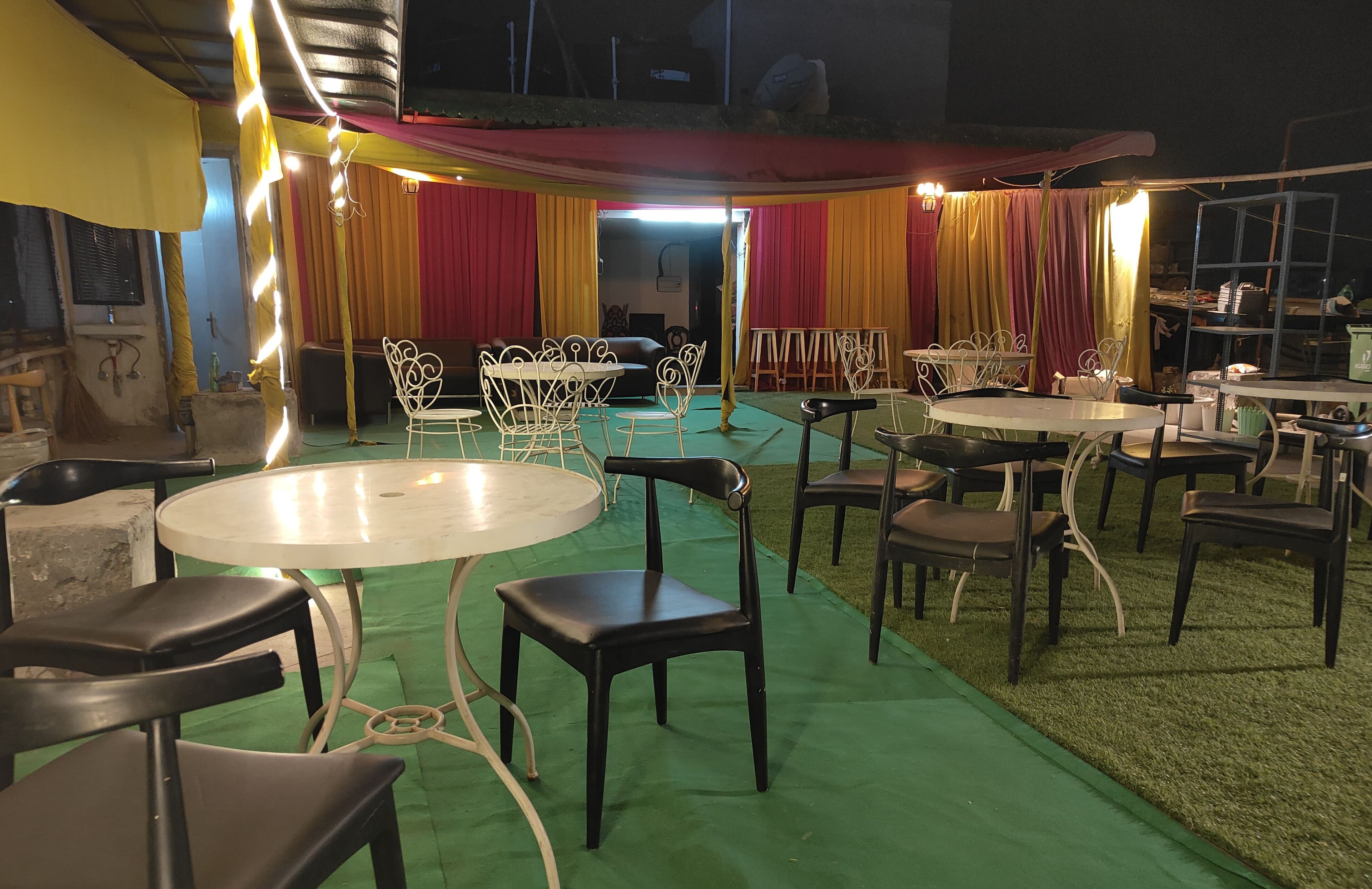 Un área de comedor al aire libre con mesas y sillas redondas, un escenario con cortinas y una alfombra verde.