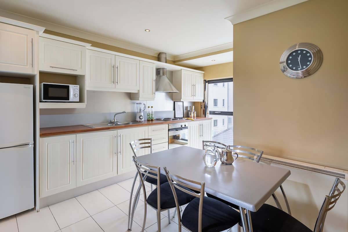 Une cuisine compacte avec des meubles blancs, un four à micro-ondes, une cuisinière et un évier. On y trouve une table à manger avec des chaises et une horloge au mur.
