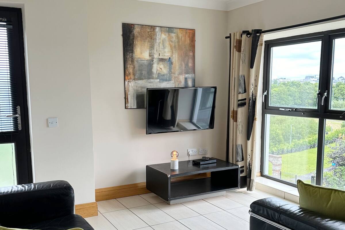Un salon moderne avec un téléviseur à écran plat fixé au mur, un canapé en cuir noir agrémenté de coussins verts et une grande fenêtre donnant sur un paysage verdoyant.