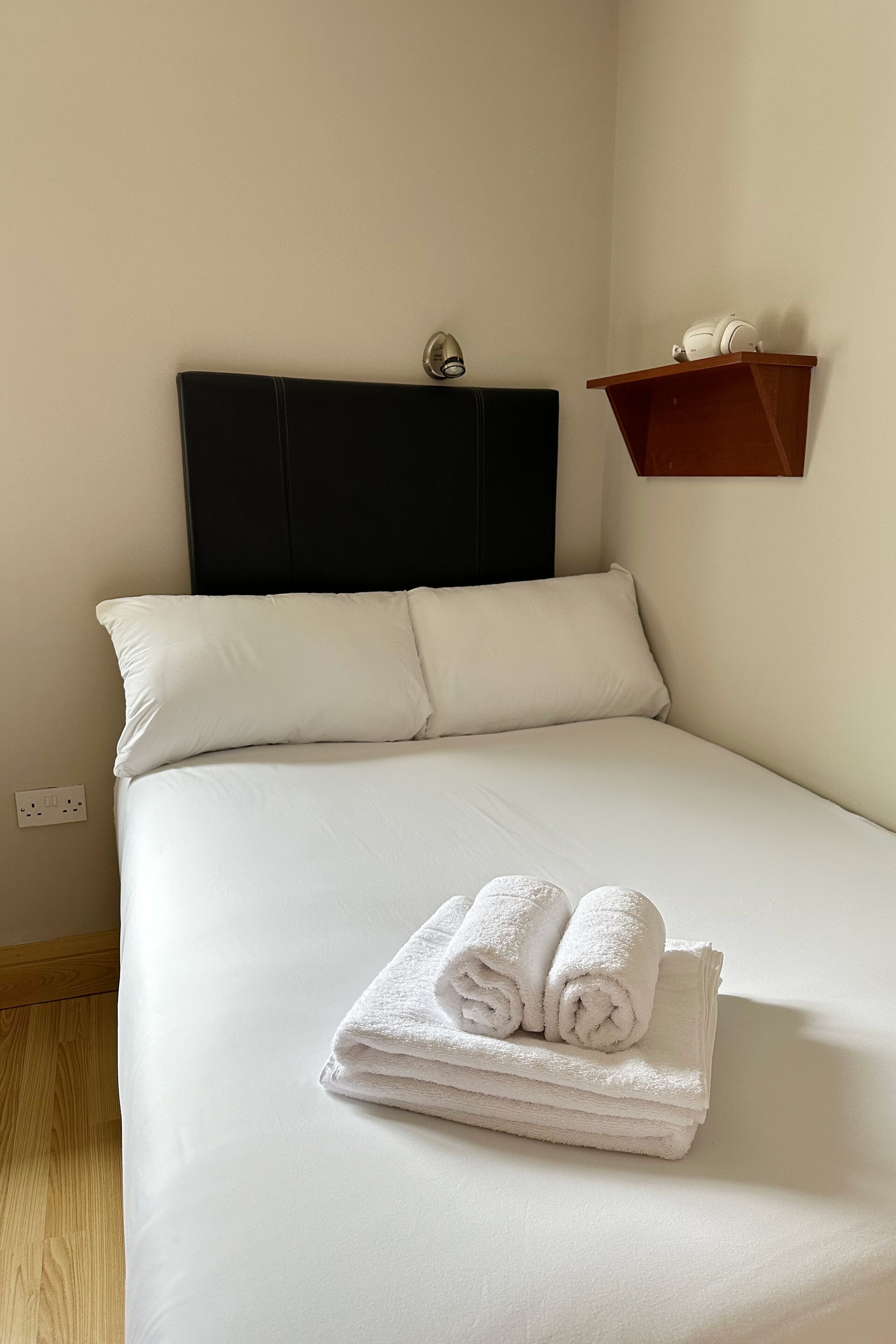 Una cama bien hecha con sábanas blancas y un cabecero, una mesita de noche de madera con una lámpara y un pequeño estante con una tetera.