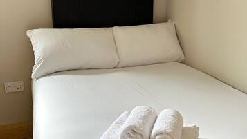 Una cama bien hecha con sábanas blancas y un cabecero, una mesita de noche de madera con una lámpara y un pequeño estante con una tetera.