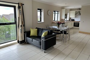 A modern living room with a black leather sofa, a dining table with chairs, and a kitchen area in the background. - Cúirt na Coiribe Self Catering (Galway)