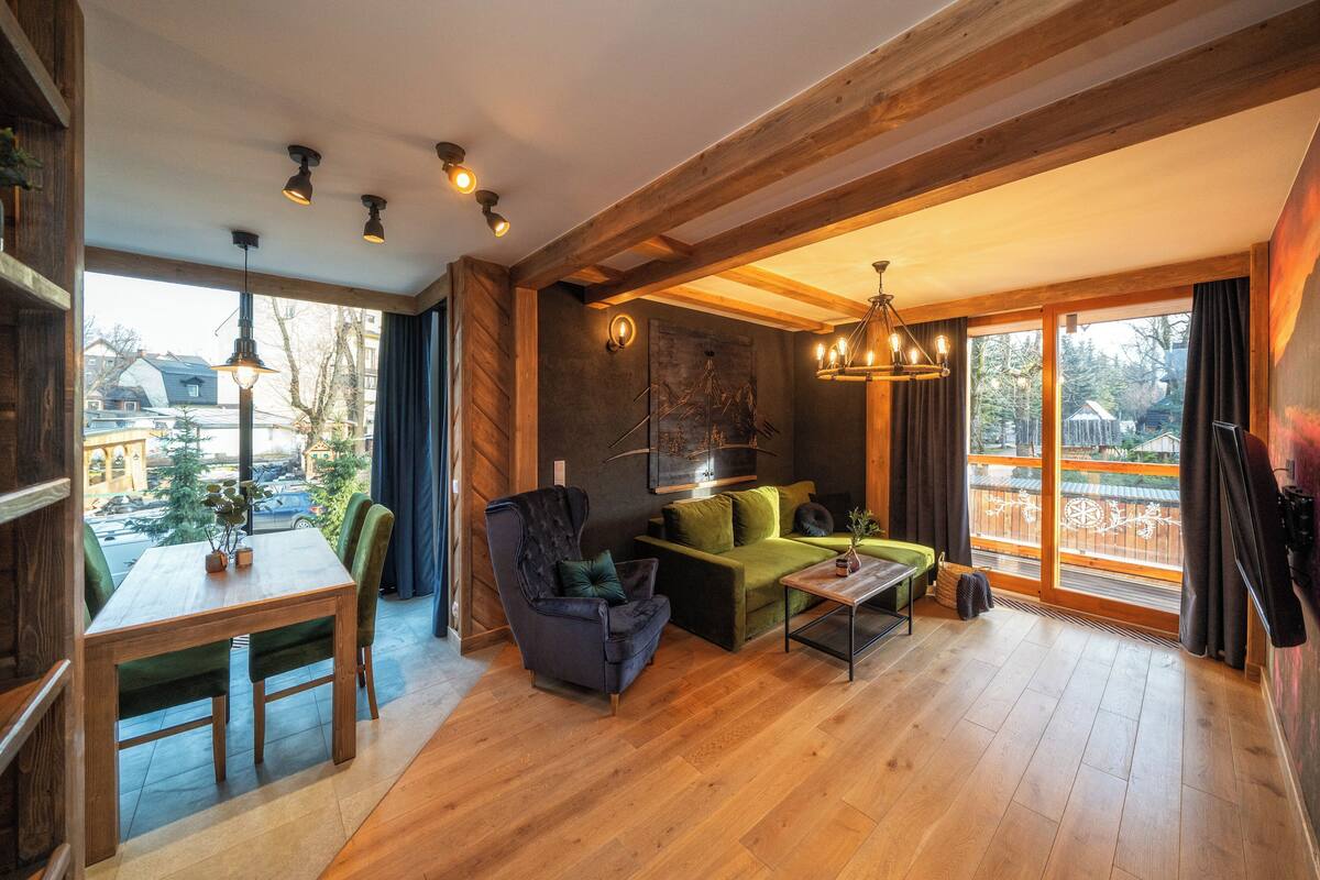A spacious living area with a wooden floor, a sofa, a chair, a table, and a large window offering a view of the outside.