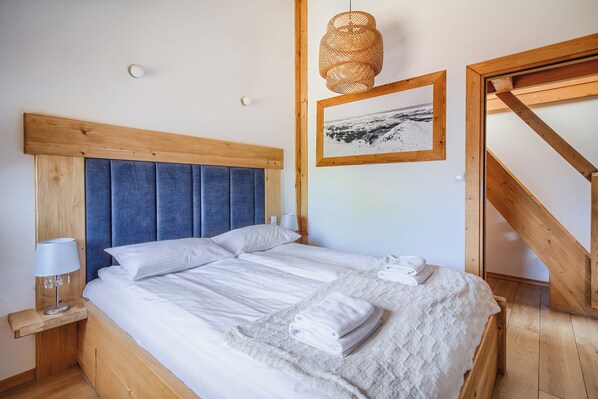 A neatly made bed with white linens and a folded towel, a wooden headboard, a wall-mounted picture, and a wooden staircase.