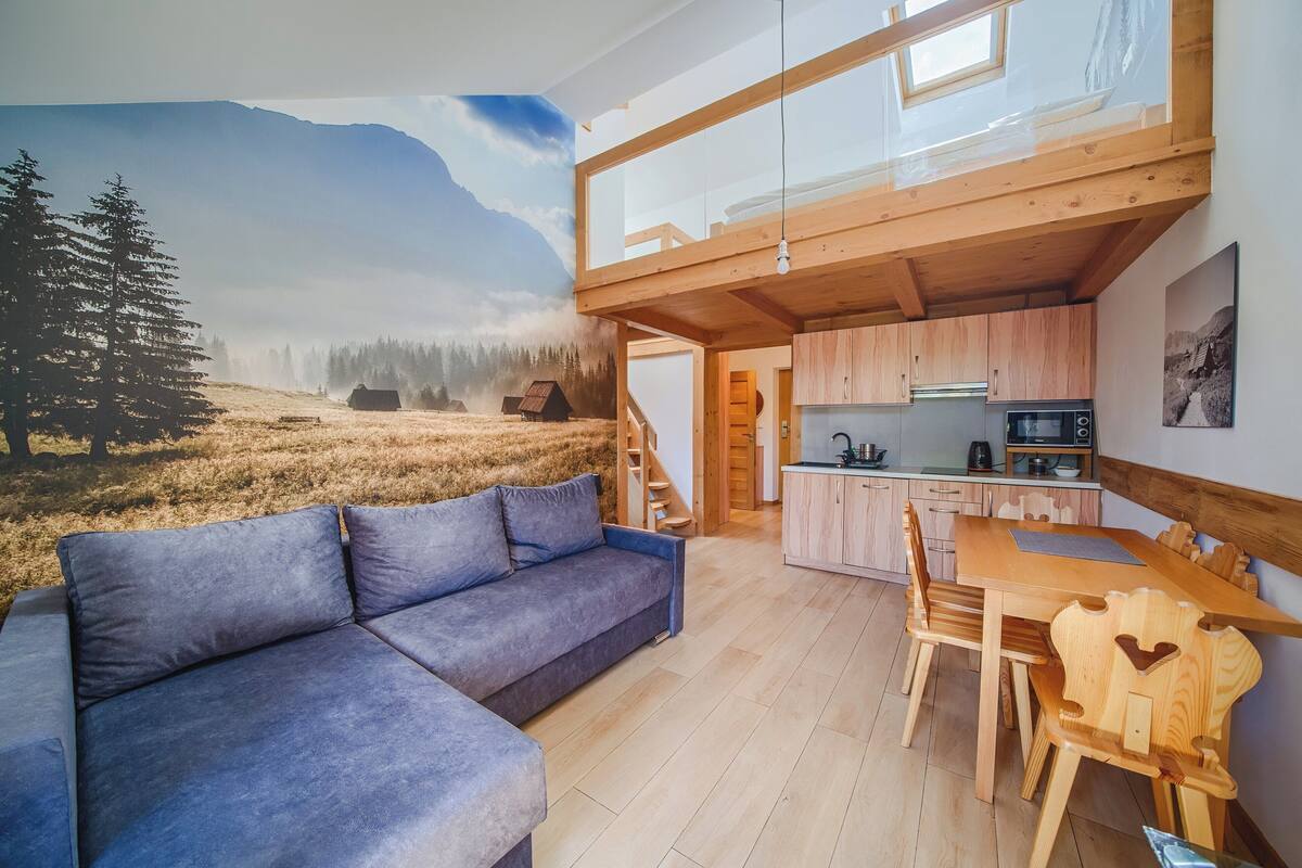 A cozy living area with a sofa, wooden dining table, and chairs. A kitchen with cabinets and appliances is visible in the background.