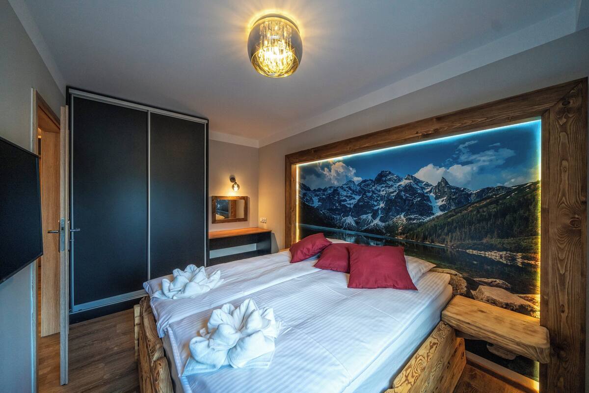 A bedroom with a large bed, a wooden headboard, a wall-mounted TV, and a mountain landscape mural.