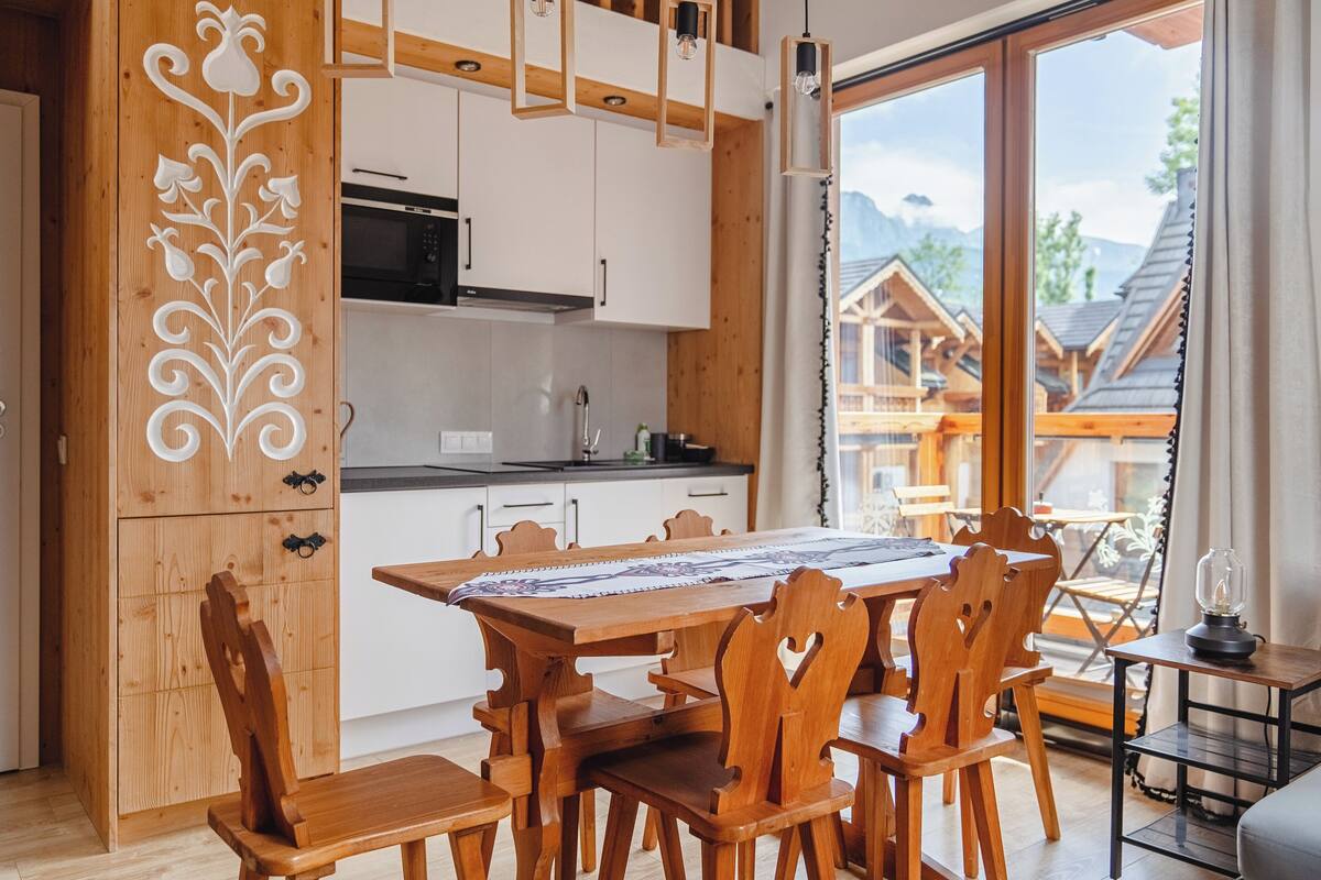 A wooden dining table with chairs, a kitchen with white cabinets, and a view of a mountain landscape through large windows.
