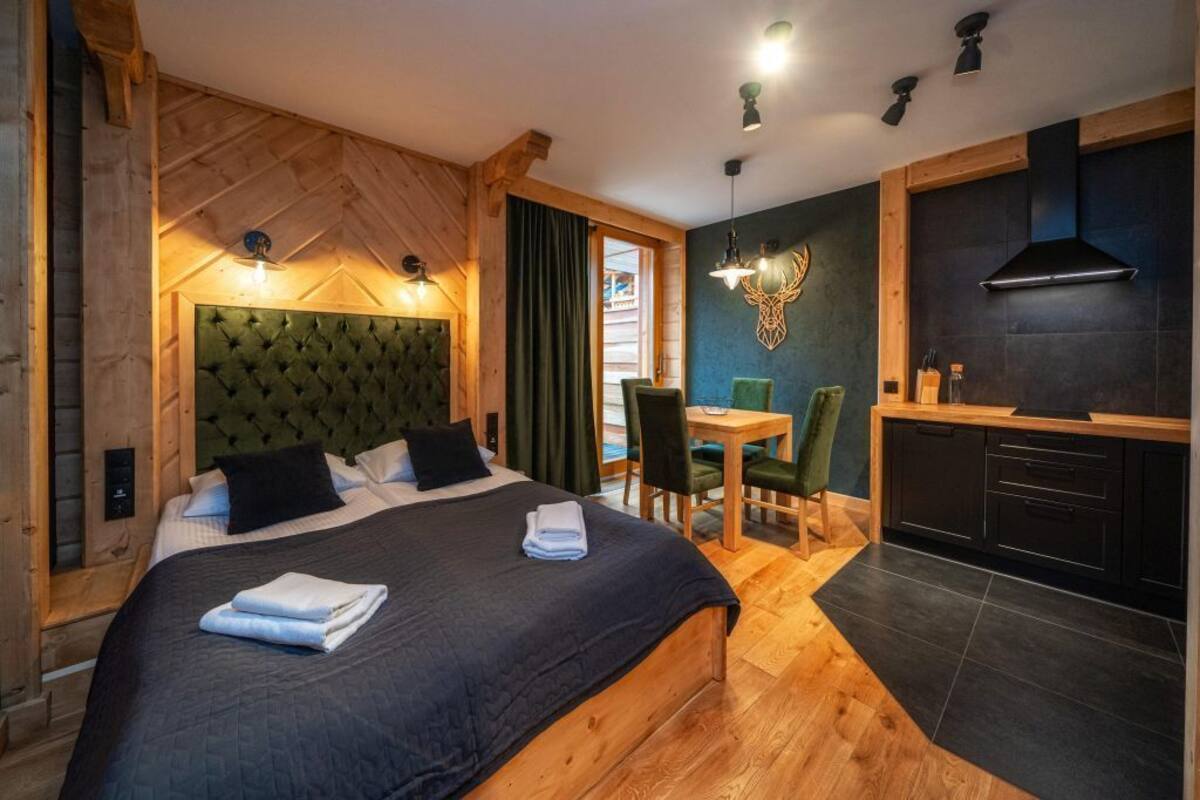 A bedroom with a large bed, a wooden headboard, a dining area with a table and chairs, and a kitchen area with a stove and cabinets.