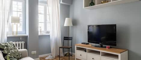 Un salon avec un téléviseur, un fauteuil, un canapé et une fenêtre avec des rideaux.