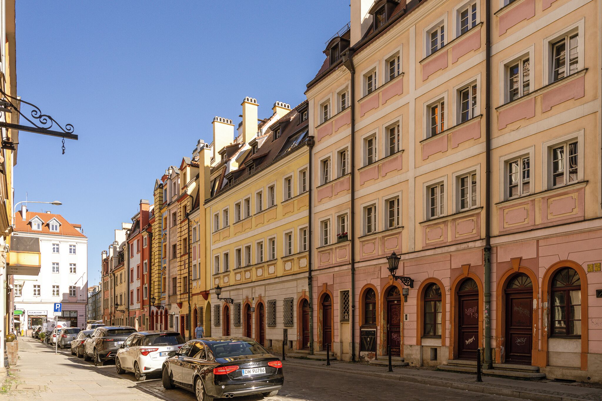 Une rangée de voitures garées le long d’une rue bordée d’immeubles à plusieurs étages, dotés d’arcs en plein cintre et de balcons.
