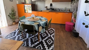 A modern kitchen with orange cabinets, a dining area with a patterned rug, and a wooden floor. - "vers L'horizon" - Maison de Vacances à 800m de Cajarc Dans le Lot (Salvagnac-Cajarc)
