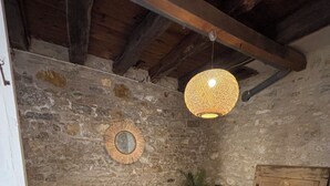 A rustic room with stone walls, a wooden ceiling, a woven light fixture, a round wooden table, a white plastic chair, a framed picture, and a potted plant. - "vers L'horizon" - Maison de Vacances à 800m de Cajarc Dans le Lot (Salvagnac-Cajarc)