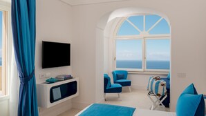 A modern hotel room with a large bed, a TV, a seating area with chairs, and a view of the sea through a window.