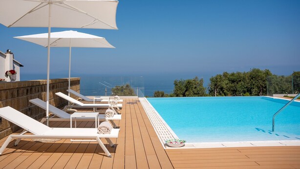 A poolside area with lounge chairs and umbrellas, a wooden deck, and a view of the sea.