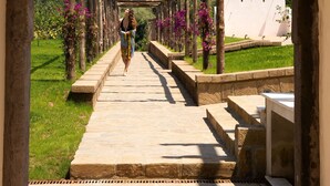 A stone pathway leading through an arched wooden structure with steps and benches, surrounded by greenery and a clear sky.