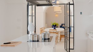 A modern kitchen with white cabinetry, a central island with pots, a hanging light fixture, and an arched doorway leading to a dining area with a table and chairs.