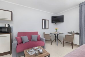 A modern hotel room with a red sofa, a small dining table with chairs, a flat-screen TV, and a kitchenette.
