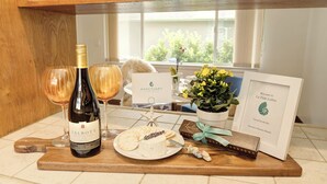 A wine bottle, two glasses, a plate with cheese and crackers, a small gift box, a framed welcome sign, and a potted plant on a wooden counter.