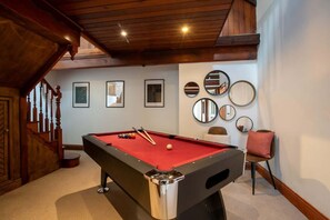 A pool table in a room with a wooden staircase, framed pictures on the wall, and a red carpet.