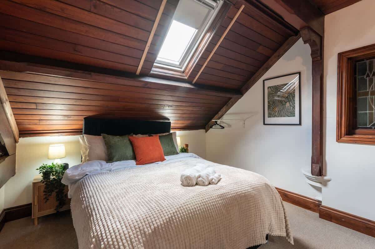 Um quarto com teto inclinado, vigas de madeira, claraboia, cama com colcha branca e uma obra de arte emoldurada na parede.