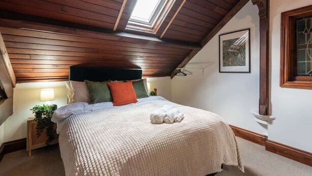 Une chambre avec un plafond en pente, des poutres en bois, un puits de lumière, un lit recouvert d’une courtepointe blanche et une œuvre d’art encadrée accrochée au mur.