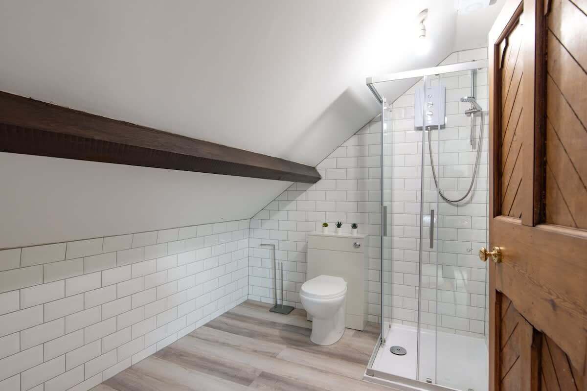 A modern bathroom with white tiled walls, a wooden beam ceiling, a glass shower enclosure, and a white toilet.