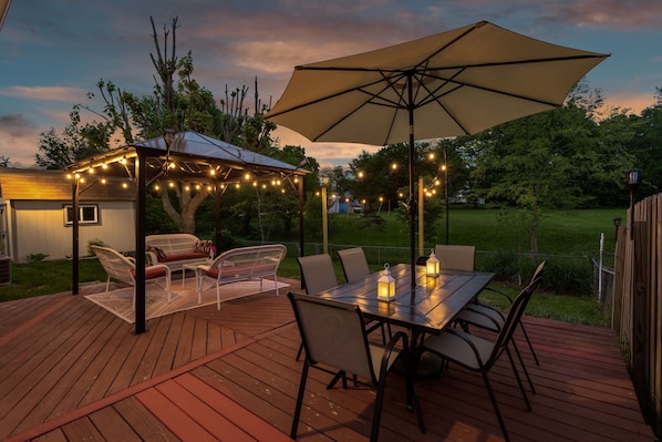 Una terraza de madera con una mesa y sillas, un cenador con luces colgantes y un paraguas grande.