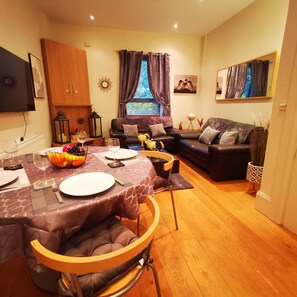 A cozy living room with a dining area, wooden flooring, and a large window with curtains.