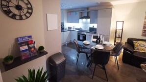 Una sala de estar moderna con zona de comedor, que incluye un gran reloj de pared, una estantería con libros y un mapa, un cubo de basura y un sofá con un cojín estampado.