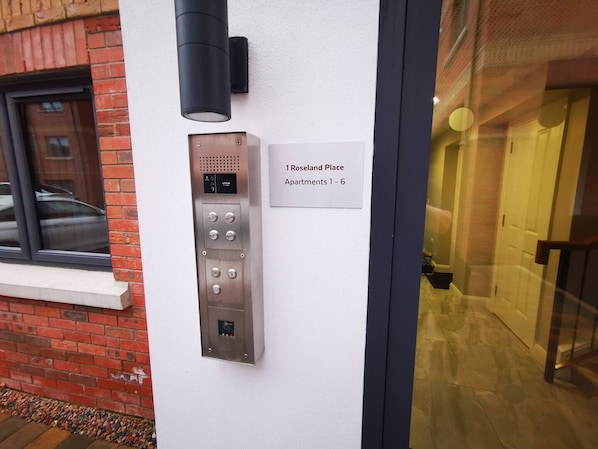 Une entrée d’immeuble avec un panneau de commande numérique pour l’ascenseur, une enseigne indiquant « 1 Roseland Place, appartements 1 à 6 », et une porte en verre donnant accès à l’intérieur.
