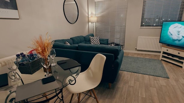 Ein modernes Wohnzimmer mit einem dunkelgrünen Sofa, einem Glastisch und einem weißen Stuhl. Rechts befindet sich ein Fernseher und links eine Lampe.