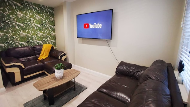 Ein Wohnzimmer mit einem braunen Ledersofa, einem Holztisch und einem an der Wand befestigten Fernseher, der das YouTube-Logo zeigt.