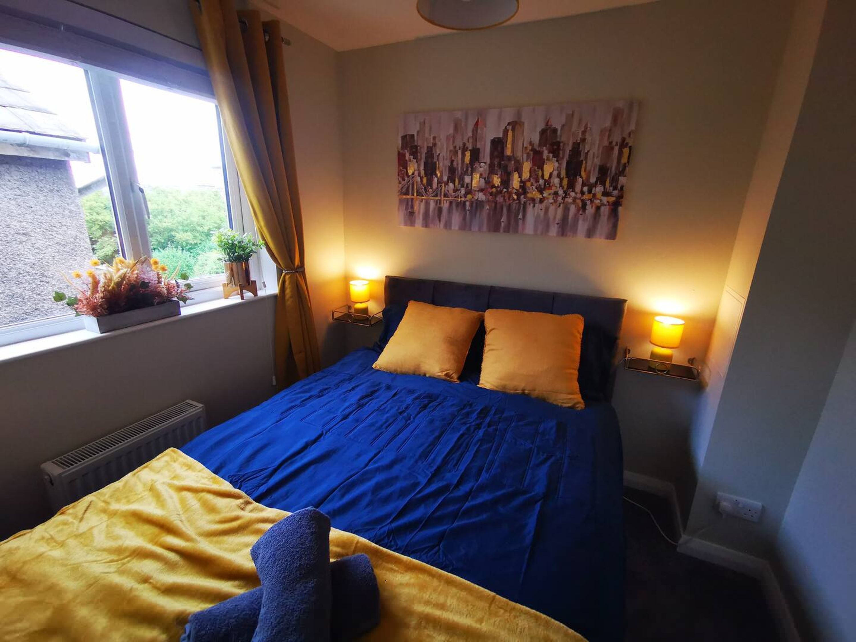 A bedroom with a bed, two bedside lamps, a window with curtains, and a wall-mounted painting.