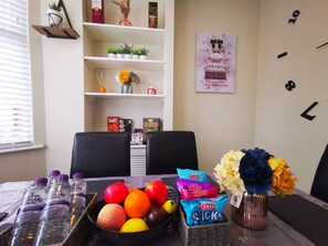 Un comedor con una mesa lista para dos personas, que incluye un frutero, agua embotellada y porciones de snacks.