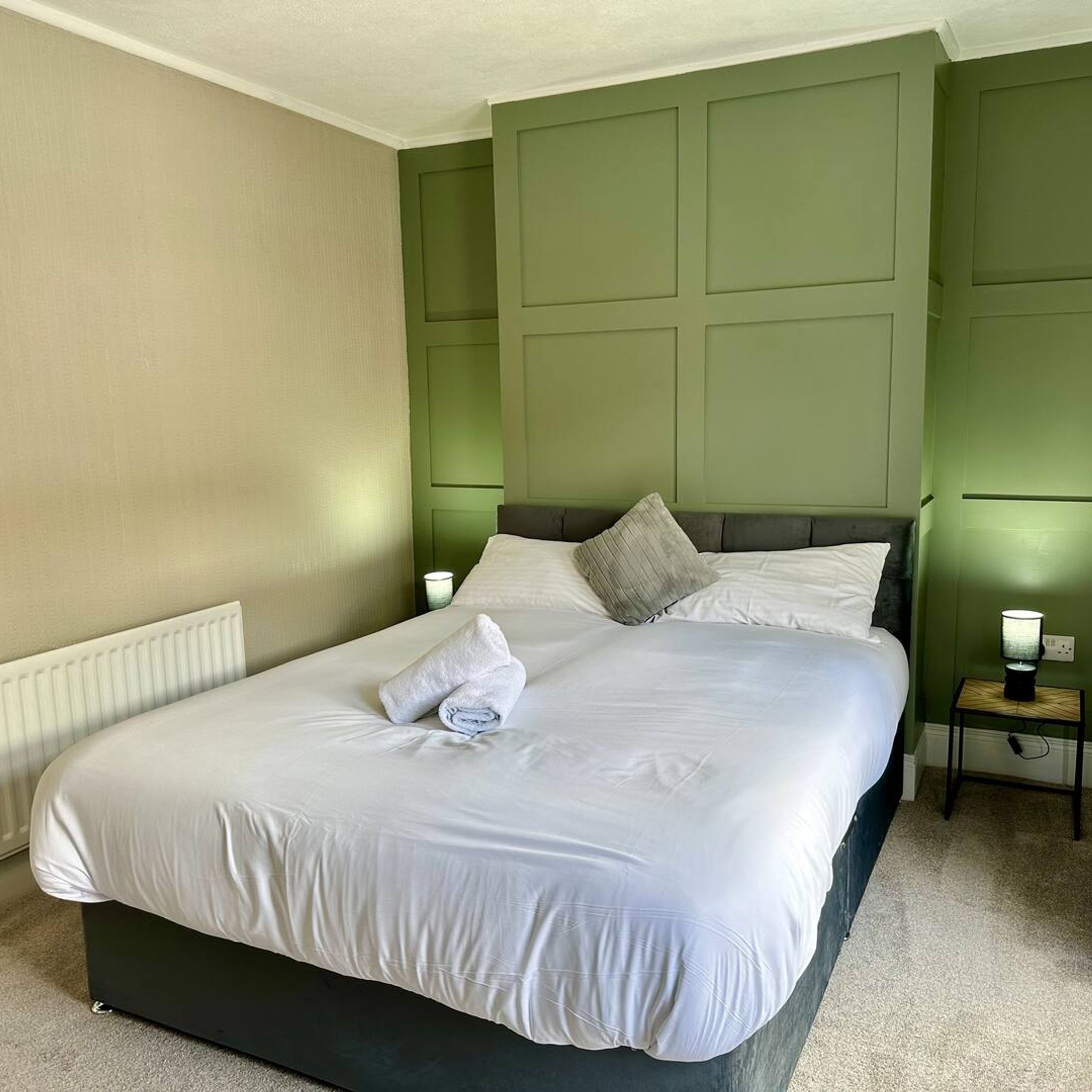 A neatly made bed with white linens and a folded towel, a bedside table with a lamp, and a green paneled wall.