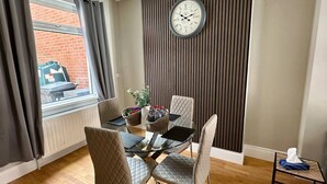 Un espace repas moderne avec une table en bois, quatre chaises, une horloge au mur et une fenêtre avec des rideaux.