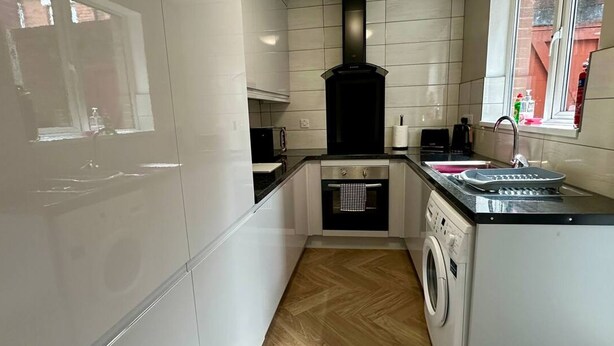 Une cuisine compacte avec des meubles blancs, une hotte noire, un lave-linge et une fenêtre donnant sur des immeubles.