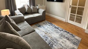 Un salon avec un canapé gris, un tapis à motifs, un téléviseur à écran plat et une porte à panneaux de verre.