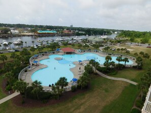A large, multi-pool outdoor swimming area with lounge chairs and umbrellas, surrounded by palm trees and a marina in the background.