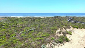 Un paisaje costero con playa de arena, vegetación verde y un paseo de madera que lleva al mar.
