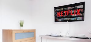 Una cocina moderna con un armario de madera, una pequeña planta en maceta, una mesa de comedor con un tazón y una copa de vino, y un televisor mostrando contenido de Netflix.
