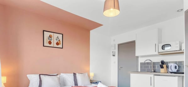 Un dormitorio con cama, almohadas, una lámpara de noche, un cuadro en la pared y una kitchenette con microondas y utensilios.